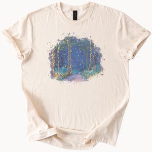 May include: Cream-colored t-shirt featuring a watercolor painting of a forest scene. The artwork depicts tall trees with a path leading through the woods, illuminated by soft yellow and blue hues, creating a dreamy aesthetic.