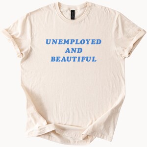 May include: Cream-colored t-shirt with the words "UNEMPLOYED AND BEAUTIFUL" printed in blue. The shirt has a crew neck and short sleeves, with the sleeves rolled up. The fabric appears soft and slightly wrinkled.