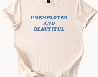 Könnte beinhalten: Cremefarbenes T-Shirt mit dem blauen Aufdruck "UNEMPLOYED AND BEAUTIFUL". Das Shirt hat einen Rundhalsausschnitt und kurze Ärmel, die hochgekrempelt sind. Der Stoff wirkt weich und leicht zerknittert.
