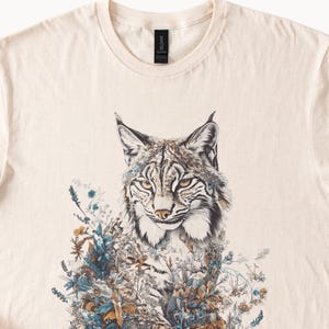 May include: Cream-colored t-shirt featuring a detailed illustration of a lynx with intricate floral and botanical elements. The lynx has a focused gaze, with a blend of brown, white, and black fur. The design incorporates shades of blue, brown, and green.