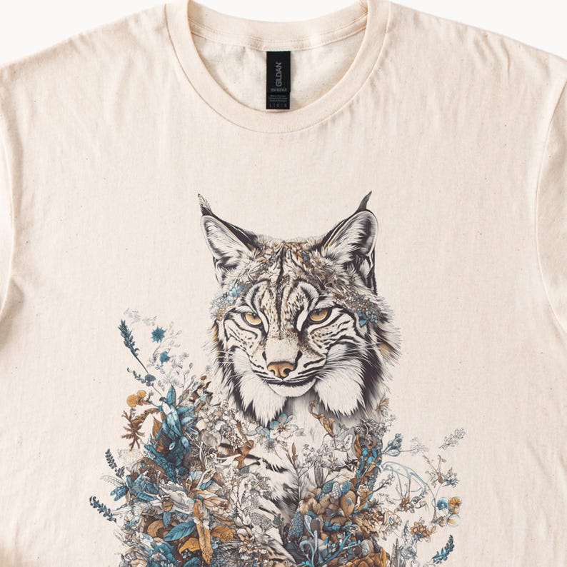 May include: Cream-colored t-shirt featuring a detailed illustration of a lynx with intricate floral and botanical elements. The lynx has a focused gaze, with a blend of brown, white, and black fur. The design incorporates shades of blue, brown, and green.