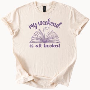 May include: Cream-colored t-shirt with the phrase "my weekend is all booked" in purple script. The design features an open book with a heart above it. The shirt has short sleeves and a crew neck.
