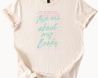 May include: A cream-coloured t-shirt featuring the teal text "Ask me about my Books". The design incorporates a pink grid pattern and the phrases "Keep this book clean" and "Do not turn down the leaves".