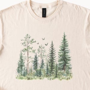May include: Cream-colored t-shirt featuring a watercolor forest scene. The design includes various green trees, including pines and deciduous trees, with small birds flying above. The shirt has a crew neck and short sleeves.