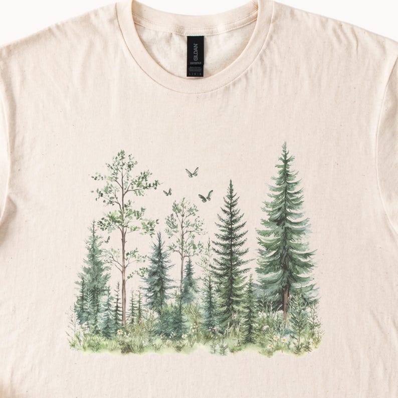 May include: Cream-colored t-shirt featuring a watercolor forest scene. The design includes various green trees, including pines and deciduous trees, with small birds flying above. The shirt has a crew neck and short sleeves.