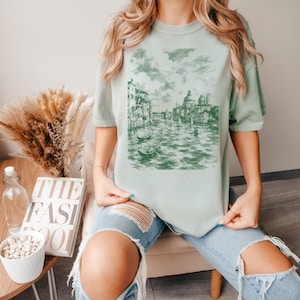 May include: A light sage green short-sleeve t-shirt featuring a dark green monochromatic sketch print of a Venetian canal scene with buildings, boats, and water. The graphic is centered on the chest of the crew neck garment.