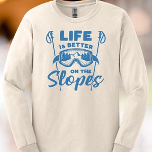 Op de afbeelding: Crèmekleurig shirt met lange mouwen en een blauwe print. De print bevat een skibril, skistokken en de tekst "LIFE is BETTER ON THE Slopes". Het shirt is gemaakt van een zacht materiaal.