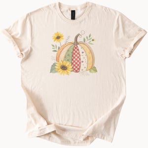 以下が含まれることがあります： クリーム色のTシャツ。カラフルなカボチャのデザインが特徴です。カボチャは、オレンジ、緑、赤のギンガムチェックなど、さまざまな模様と色で構成されています。2つのヒマワリと緑の葉がカボチャを囲み、秋をテーマにしたデザインになっています。
