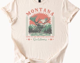 May include: A beige t-shirt with a vintage-style graphic design featuring a moose in a mountain landscape. The text "Montana" is in a retro font above the moose, and the text "Going to the Mountain Adventure Outdoors" is below the moose.