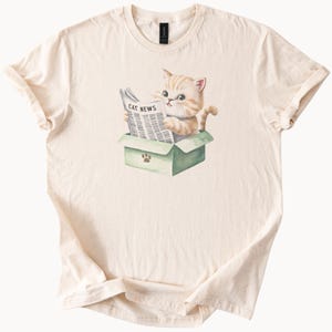 以下が含まれることがあります： クリーム色のTシャツ。水彩画で描かれたトラ猫が、緑色の箱の中で「CAT NEWS」と書かれた新聞を読んでいるイラスト。箱には足跡が描かれています。