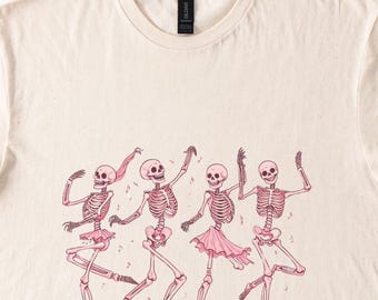 Puede incluir: Una camiseta beige con un gráfico de cuatro esqueletos bailando en tutús rosas. Los esqueletos llevan tutús rosas y tienen detalles rosas en sus huesos. El fondo es beige claro.