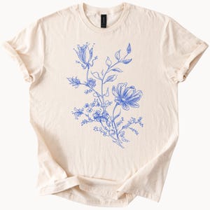 May include: Cream-colored t-shirt featuring a blue floral design. The graphic depicts a detailed botanical illustration of flowers, leaves, and stems. The t-shirt has a classic crew neck and short sleeves, perfect for casual wear.