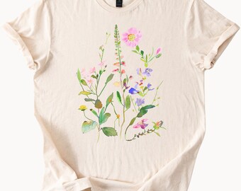 Op de afbeelding: Crèmekleurig T-shirt met een aquarel bloemenontwerp. Het kunstwerk toont verschillende bloemen in roze, paarse en gele tinten, met groene bladeren en stelen. Het shirt heeft een klassieke ronde hals en korte mouwen.