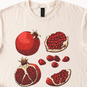 May include: Cream-colored t-shirt featuring a detailed illustration of pomegranates. The design includes a whole pomegranate, a halved pomegranate, and several pomegranate seeds. The artwork is in shades of red and brown.