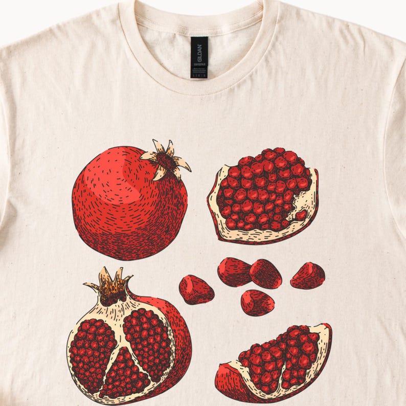 May include: Cream-colored t-shirt featuring a detailed illustration of pomegranates. The design includes a whole pomegranate, a halved pomegranate, and several pomegranate seeds. The artwork is in shades of red and brown.