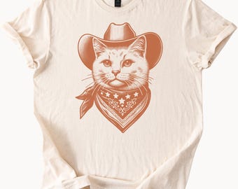 May include: A beige t-shirt with a brown illustration of a cat wearing a cowboy hat and bandana.