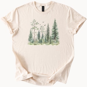 May include: Cream-colored t-shirt featuring a watercolor illustration of a forest scene. The design includes various green trees, grass, and small flying birds. The shirt is made of soft material and has a classic crew neck.