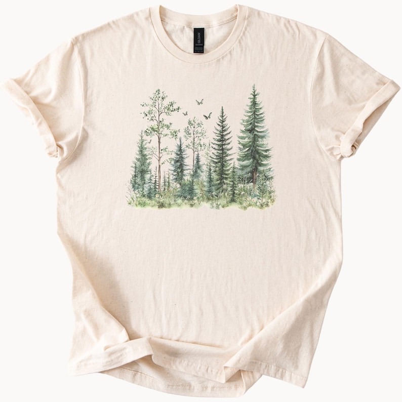 May include: Cream-colored t-shirt featuring a watercolor illustration of a forest scene. The design includes various green trees, grass, and small flying birds. The shirt is made of soft material and has a classic crew neck.