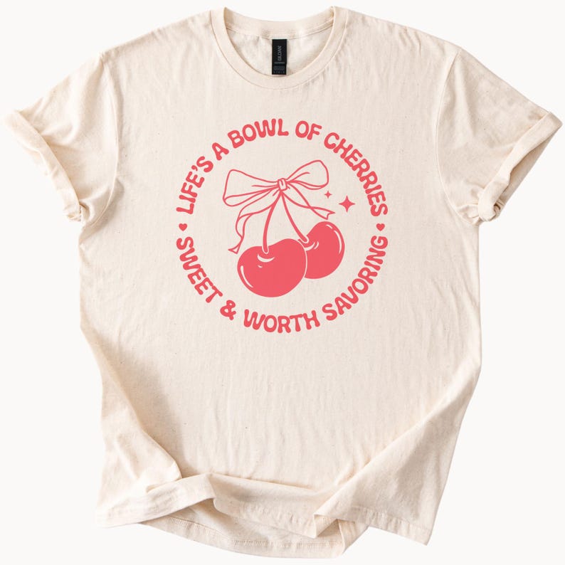 May include: A cream-colored t-shirt with a pink circle graphic that says "Life's a bowl of cherries, sweet & worth savoring". The graphic also features two red cherries with a pink bow.