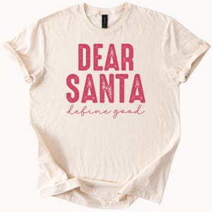 May include: A cream-coloured t-shirt featuring the text "DEAR SANTA" in a distressed pink font, with "define good" written below in a script font. The shirt has short sleeves and a crew neck.