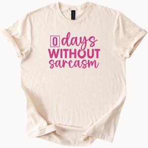 May include: Cream-colored t-shirt with the phrase "0 days WITHOUT sarcasm" in bright pink lettering. The number "0" is enclosed in a pink square. The shirt has short sleeves and a crew neck.