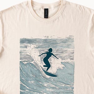 May include: A cream colored t-shirt with a blue silhouette of a person surfing on a wave. The wave is depicted in a blue and white woodblock style.