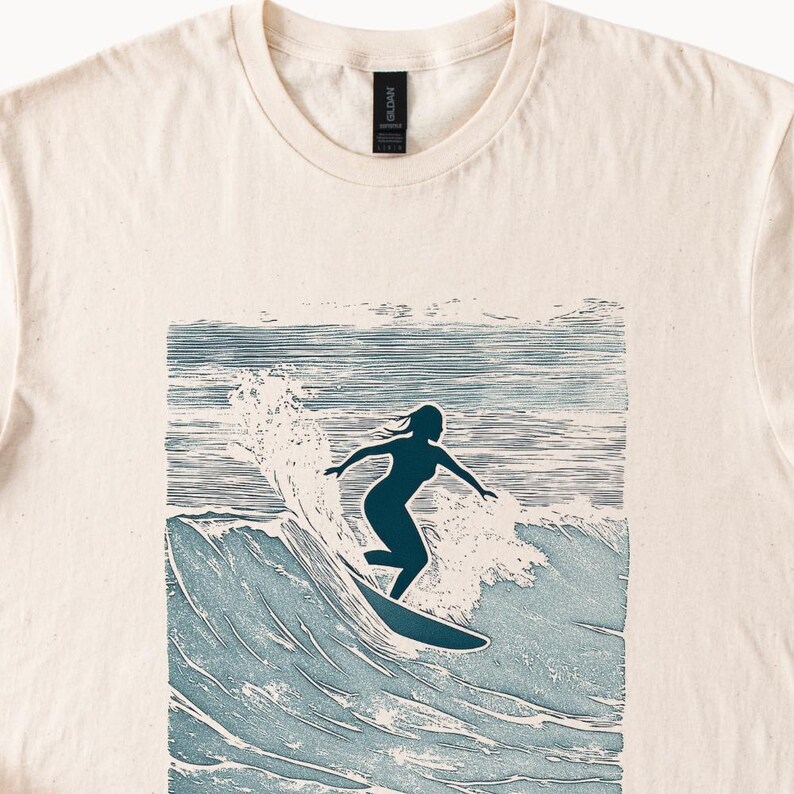 May include: A cream colored t-shirt with a blue silhouette of a person surfing on a wave. The wave is depicted in a blue and white woodblock style.