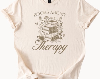 Może przedstawiać: Kremowa koszulka z napisem "BOOKS ARE MY Therapy" w brązowym skrypcie. Projekt przedstawia stos książek, filiżankę kawy, motyle i elementy kwiatowe.