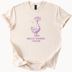 Puede incluir: Una camiseta beige con una ilustraci&oacute;n morada de un ganso con gafas y el texto "Silly Goose Club."
