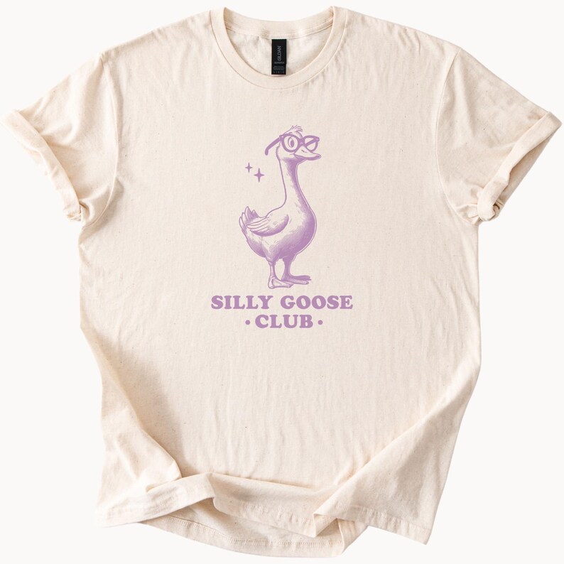 Puede incluir: Una camiseta beige con una ilustraci&oacute;n morada de un ganso con gafas y el texto "Silly Goose Club."