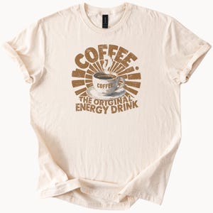 以下が含まれることがあります： クリーム色のTシャツに茶色のグラフィックデザイン。デザインは、コーヒーカップの画像の上に「COFFEE」の文字をあしらっています。カップの下には「THE ORIGINAL ENERGY DRINK」と書かれています。レトロでヴィンテージな雰囲気のデザインです。