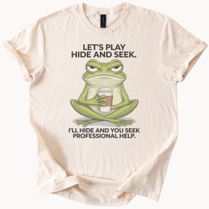 Puede incluir: Una camiseta beige con un dibujo animado de una rana verde sosteniendo una taza de café. El texto en la camiseta dice "Let's play hide and seek. I'll hide and you seek professional help."