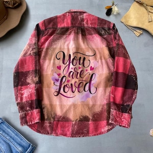 Könnte beinhalten: Rotes und schwarzes kariertes Flanellhemd mit Bleich-Effekt und den Worten "You are loved" in lila Schrift mit rosa Herzen.