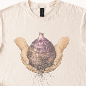 May include: A cream-colored t-shirt featuring a graphic illustration of two hands cupping a purple, bulbous root vegetable with visible roots extending from the bottom. The design is centered on the chest, showcasing detailed line work and shading.