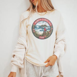 May include: Cream-colored long-sleeve shirt featuring a circular graphic with the text "North Carolina" and the year "1789". The graphic depicts a lighthouse, waves, a red bird, and foliage. The shirt is made of a soft material.