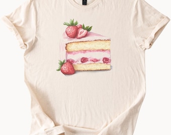 May include: A beige t-shirt featuring a graphic of a slice of strawberry cake. The cake is decorated with whipped cream and strawberries.