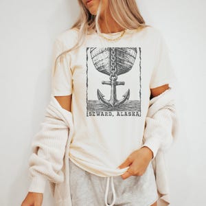 May include: A cream-colored short-sleeve t-shirt featuring a black graphic print of a ship hull connected by a chain to a large anchor. Below the anchor, the text reads SEWARD, ALASKA. The shirt is worn with a cream-colored knit cardigan.