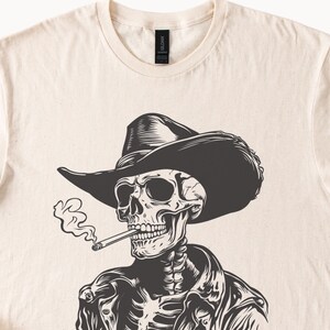 May include: A cream-coloured t-shirt with a black and white illustration of a skeleton in a cowboy hat and jacket, smoking a cigarette. The graphic is centred on the shirt, with smoke rising from the cigarette.