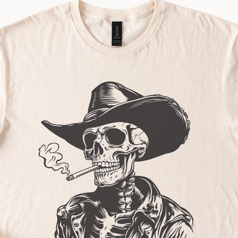 May include: A cream-coloured t-shirt with a black and white illustration of a skeleton in a cowboy hat and jacket, smoking a cigarette. The graphic is centred on the shirt, with smoke rising from the cigarette.