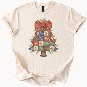 May include: Cream-colored t-shirt featuring a Christmas tree design. The tree is composed of patchwork squares with festive patterns, including snowflakes, birds, and deer. A large red bow with gold polka dots and a star adorn the top of the tree.
