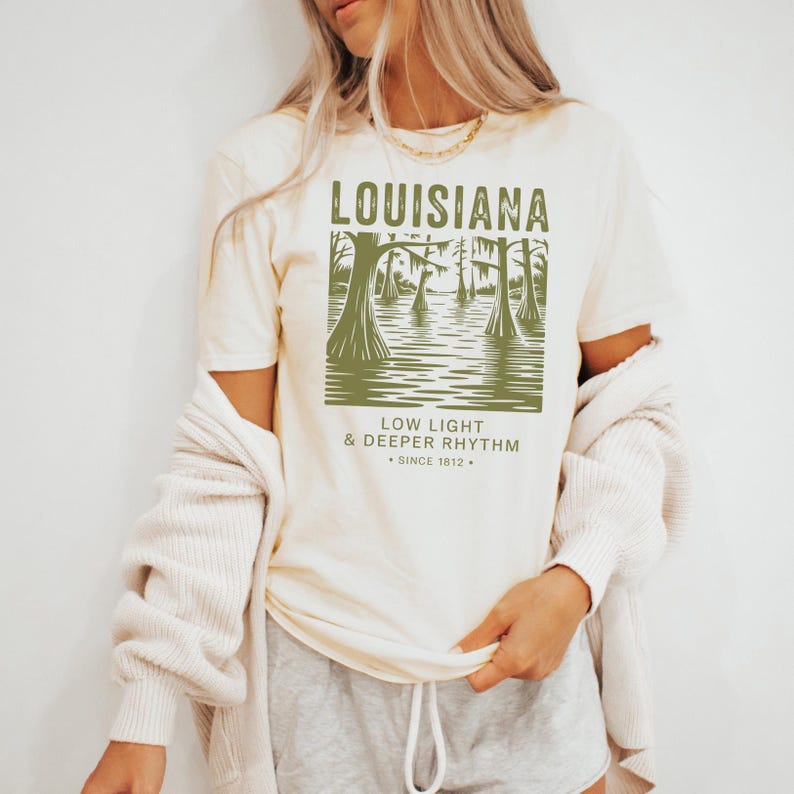 May include: Cream-colored t-shirt featuring a green graphic of a Louisiana swamp scene with text that reads "LOUISIANA, LOW LIGHT & DEEPER RHYTHM, SINCE 1812." The shirt has short sleeves and a relaxed fit.