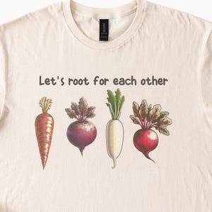 May include: A beige t-shirt with the text "Let's root for each other" printed on the front. The text is surrounded by illustrations of a carrot, a beet, a radish, and a turnip.