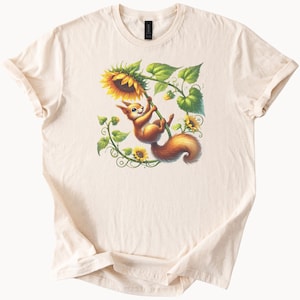 May include: Cream-colored t-shirt featuring a whimsical illustration of a squirrel swinging from a sunflower. The squirrel is depicted with orange fur and a fluffy tail. The sunflower has yellow petals and green leaves.