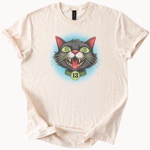 May include: A beige t-shirt with a black cat face graphic. The cat has green eyes, a red tongue sticking out, and a green collar with the number 13 on it.