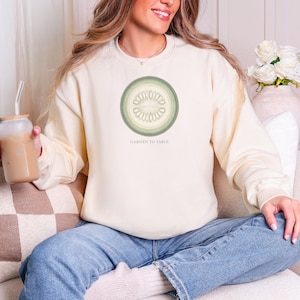 May include: A cream-colored crewneck sweatshirt featuring a circular graphic of a sliced cucumber in shades of sage and olive green. Below the graphic, the text GARDEN TO TABLE is printed in a simple, clean, sans-serif font on the chest area.