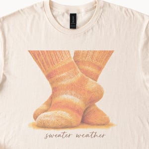 May include: A cream-colored crew neck t-shirt featuring a graphic illustration of two crossed knitted socks in shades of orange, yellow, and white. Below the socks, the text "sweater weather" is printed in a brown, cursive script font.