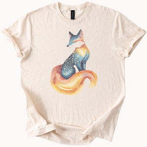 May include: A beige t-shirt with a colorful fox illustration. The fox has a blue and purple body with a constellation pattern and a long orange and yellow tail.