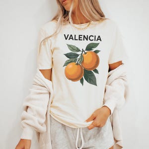 May include: A white t-shirt with a graphic of oranges on a branch and the word "VALENCIA" printed in black.
