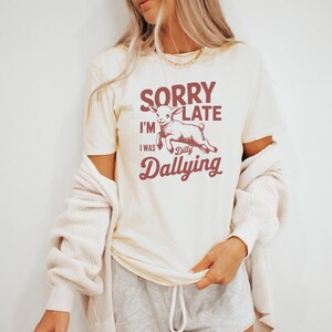 May include: Cream-colored t-shirt with the text "SORRY I'M LATE I WAS Dilly Dallying" in burgundy. A cartoon goat is featured in the center. The shirt has short sleeves and a crew neck.