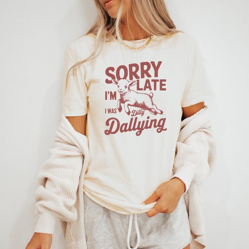 May include: Cream-colored t-shirt with the text "SORRY I'M LATE I WAS Dilly Dallying" in burgundy. A cartoon goat is featured in the center. The shirt has short sleeves and a crew neck.
